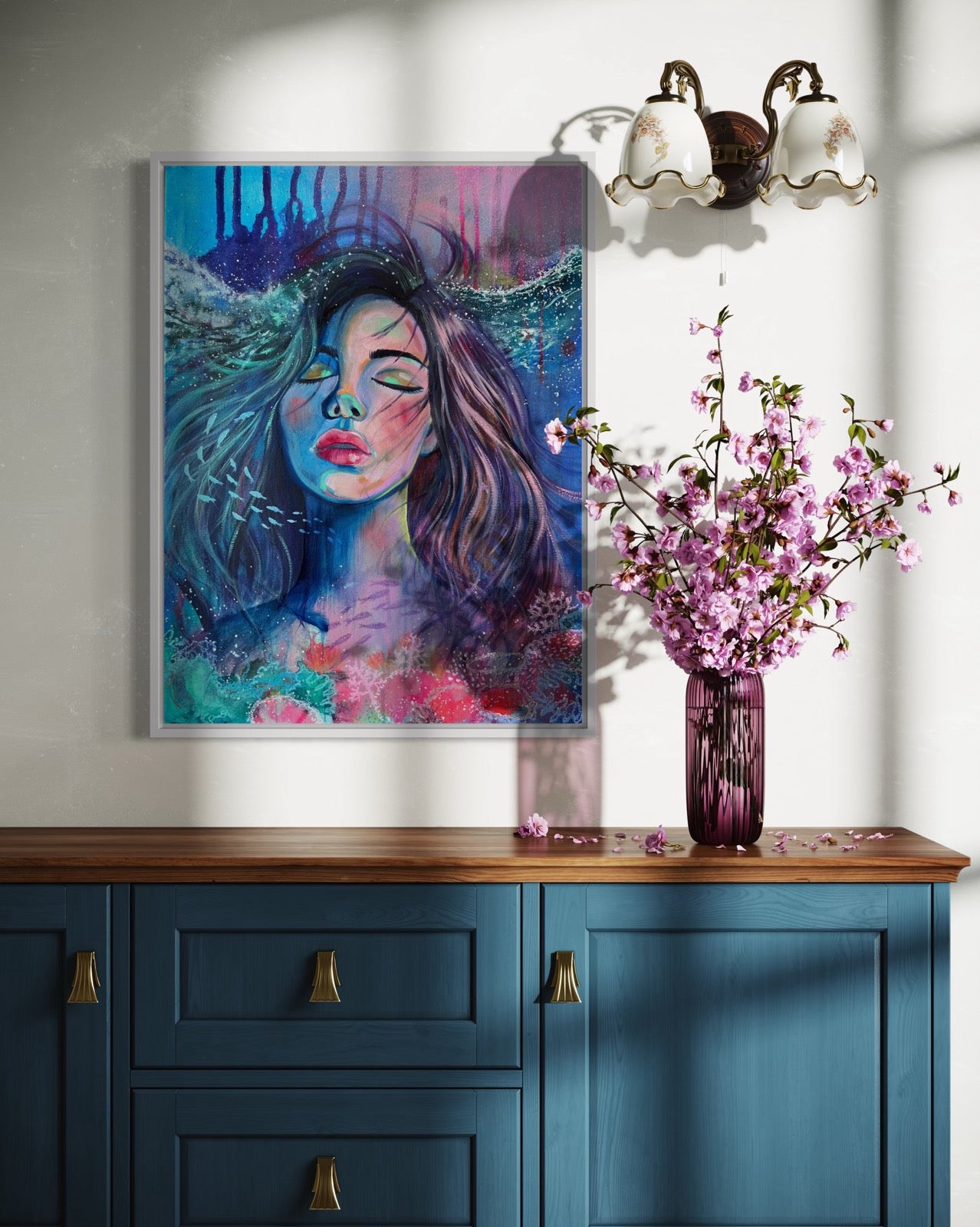 Blue and modern portrait artwork of a woman with long hair in an underwater setting a wall above a blue cabinet with a vase of pink flowers.