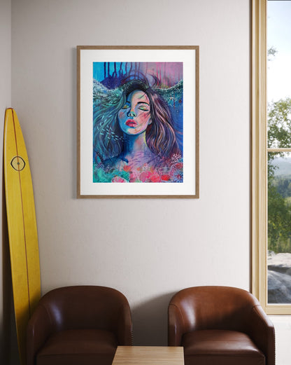 Framed blue and modern portrait artwork of a woman with long hair in an underwater setting on a white wall with beach decor on a wall above two brown chairs with yellow surf board decor